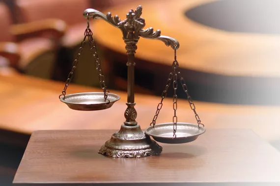 An ornately carved scale of justice rests on a table in the foreground. In the background are chairs and a circular table out of focus.