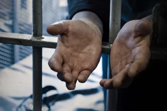 Two open hands extend through metal prison bars. The palms face upward, showing lined, calloused and weathered skin