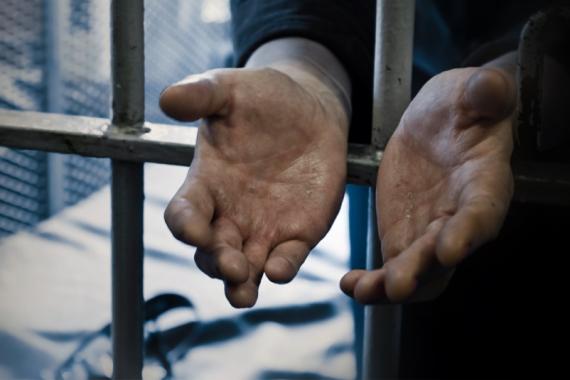Two open hands extend through metal prison bars. The palms face upward, showing lined, calloused and weathered skin