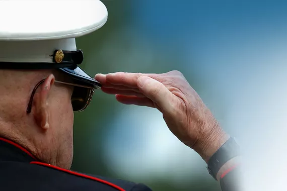 A view of a man’s head from the back. The man wears a white military hat with a dark brim. He is saluting with his right hand