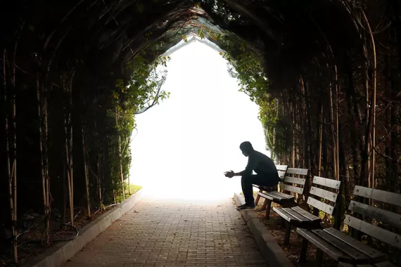 A person sits alone on a bench at the end of a dark stone tunnel. Ivy grows on the tunnel walls. The tunnel opens to a lighted area