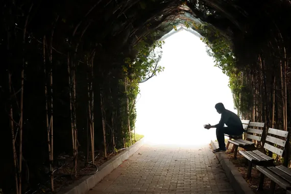 A person sits alone on a bench at the end of a dark stone tunnel. Ivy grows on the tunnel walls. The tunnel opens to a lighted area