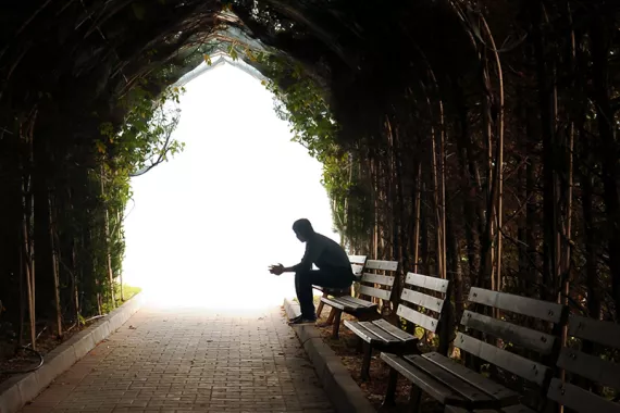 A person sits alone on a bench at the end of a dark stone tunnel. Ivy grows on the tunnel walls. The tunnel opens to a lighted area