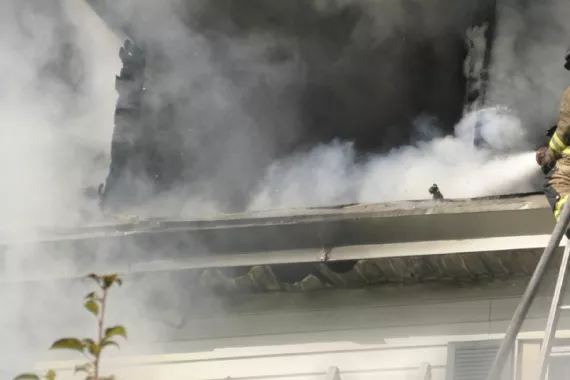 Smoke from a fire surrounds a structure as a firefighter on a ladder hoses down the roof.