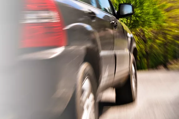 A black car drives down a road with trees in the background. A blur effect on the photo implies the car is driving at a high speed.