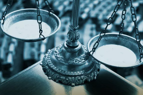 Scales of Justice with an elaborate base and two trays slightly unbalanced. Rows of courtroom chairs fill the background in shades of blue