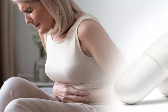 A sitting woman has her hands over her stomach. She is doubled over and in pain. An enlarged picture of a white capsule is beside her image