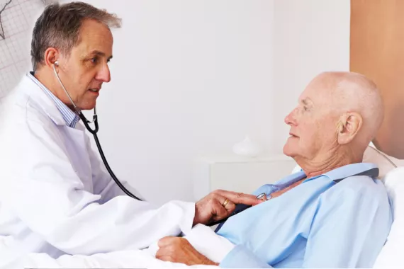 A doctor and a patient are in a hospital room. The patient is lying down and the doctor is holding a stethoscope on the patient’s chest