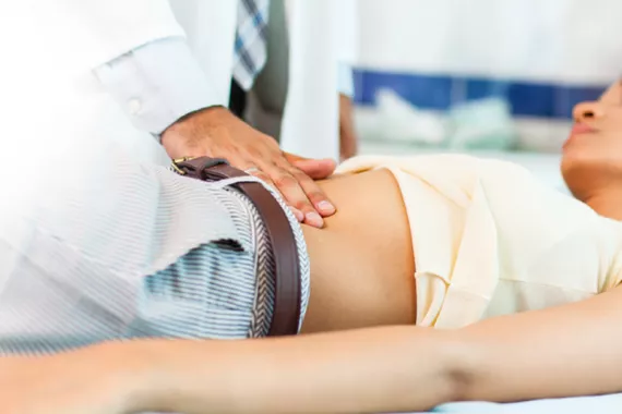 A doctor examines a young female patient lying on an exam table in a doctor’s office. The doctor is examining the patient’s abdomen