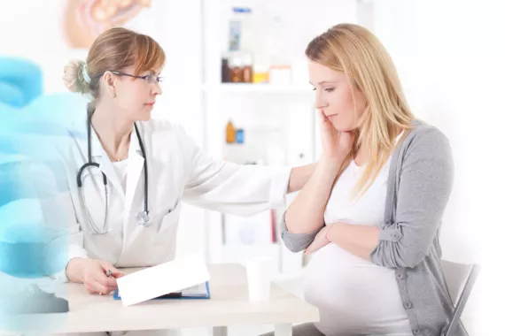 A female doctor touches the shoulder of a pregnant woman as they sit at a desk. They both have serious expressions on their faces.