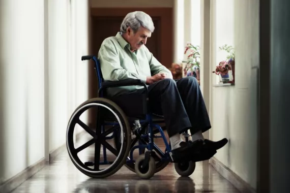 In a hallway with windows, an older man in a wheelchair looks down. Potted flower plants rest on the windowsills.