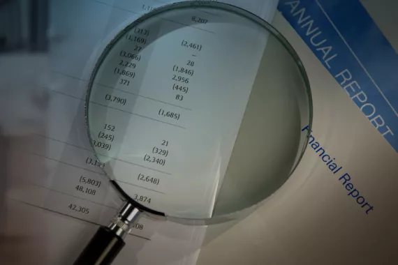 A magnifying glass rests on a short stack of papers on a desk, magnifying a budget report.