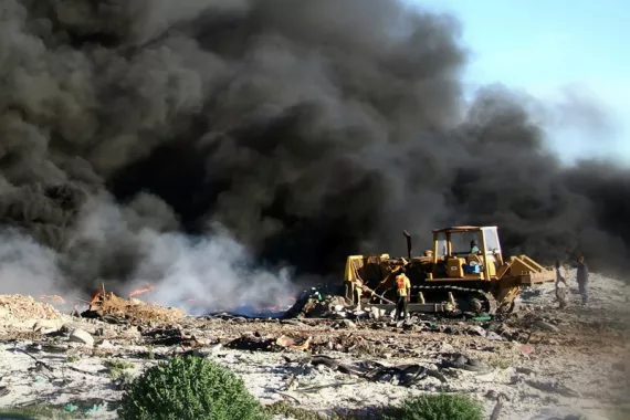 An open-air burn pit surrounded by trash and debris. Thick black smoke from the burn fills the sky. A bulldozer pushes trash into the smoke