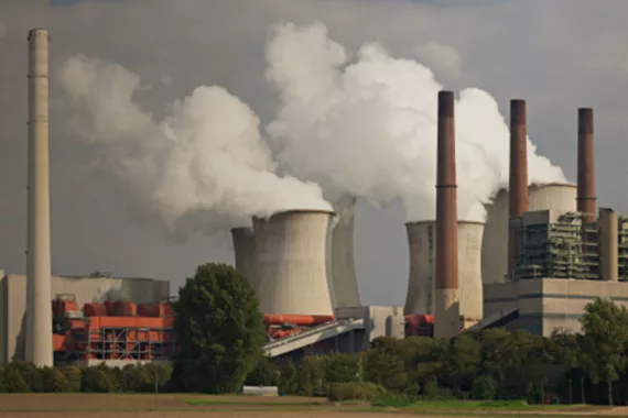 Industrial site with seven smokestacks. Four are tall and thin and three are large, wide and cylindrical. White smoke gushes into the air