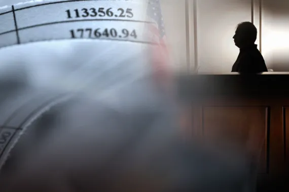 A silhouette of a man siting in a witness box in a courtroom. Paper with numbers are displayed on the wall in front of the man