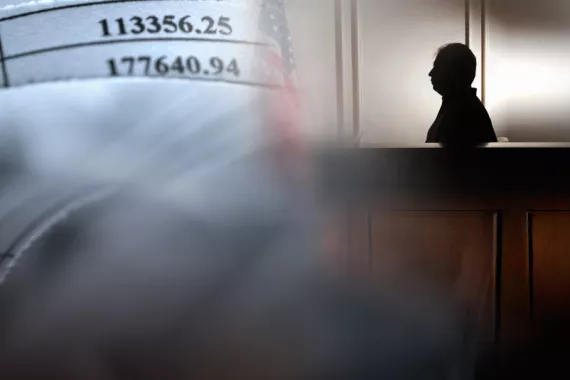 A silhouette of a man siting in a witness box in a courtroom. Paper with numbers are displayed on the wall in front of the man