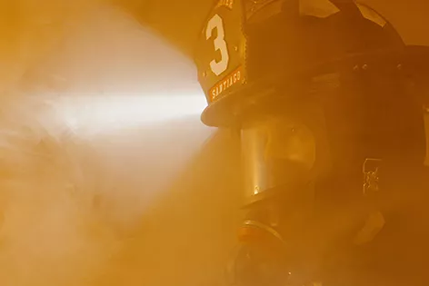 A firefighter in full protective gear working in heavy orange smoke. The helmet encloses his head and includes safety breathing apparatus