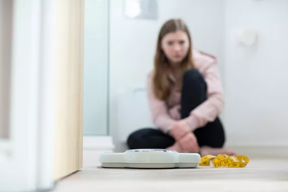 In the foreground, a scale rests on the floor next to measuring tape. In the background a young girl sits on the floor looking at the scale.