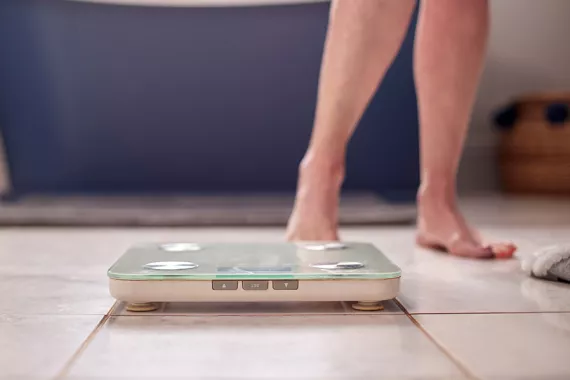 A digital scale rests on the floor in the foreground. In the background a pair of legs indicates a person is about to step onto the scale 