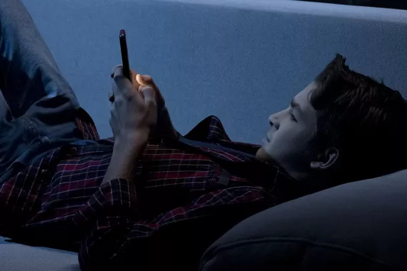 A boy lies down with his head propped up by a pillow and his phone in his hands.