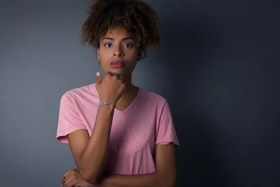 A young Black woman stands facing forward in front of a grey wall and looks straight ahead.