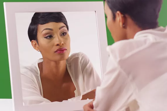 A young Black woman looks into a mirror. She wears a full face of makeup. The background in solid green.