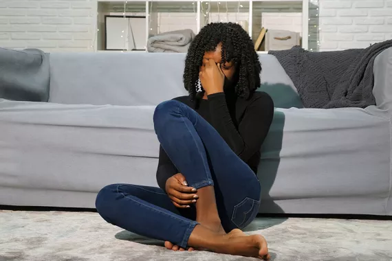 A young Black woman sits on the floor in front of a grey couch. Her hand is on her face. She looks as if she just received bad news