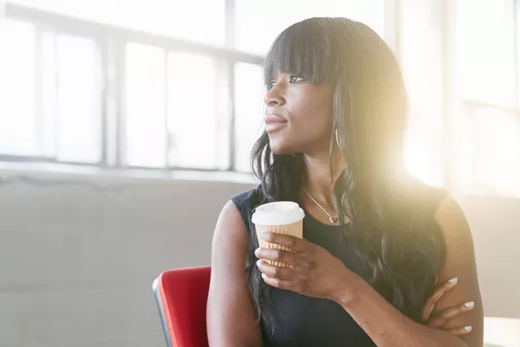 A young Black woman with long, smooth, slightly wavy hair faces left toward brightly lit windows. She holds a paper coffee cup.