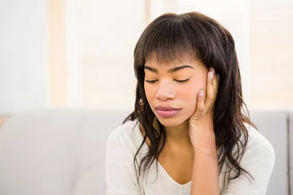 A young Black woman looking downward. The background is grey and white. Her hand holds the side of her face and she looks sad and concerned