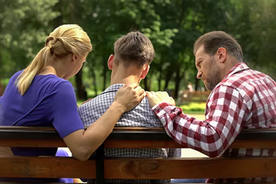 Seen from behind, a concerned man and woman sit on a bench on opposite sides of a young boy, resting their hands on his shoulders.