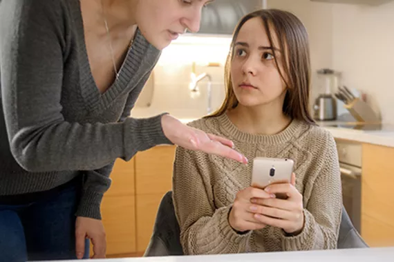 A girl holding her phone looks concerningly at an unhappy older woman gesturing to the phone.