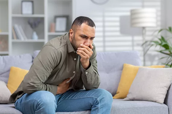 A man wearing jeans and a green button up shit holds his stomach with his right hand and covers his mouth with his left hand, indicating he is ill