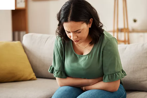 A young woman sits on a couch, leaning forward with both arms crossed over her stomach. She is frowning and looks like she is in pain