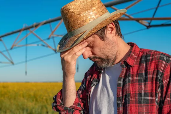 A man stands in front of a field filled with plants and a watering system overhead. The man looks downward with a worried expression