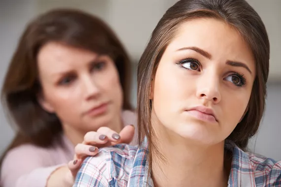 A teen girl in a blue shirt looks to the side with a serious expression. A woman behind her looks concerned and reaches out to comfort her
