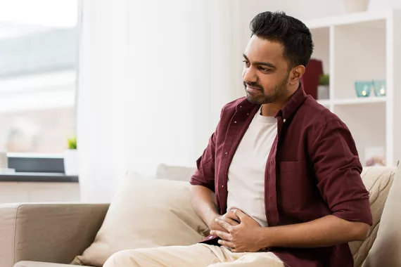 A young man is seated on a couch. Both his hands are on his stomach and looks like he is nauseated and uncomfortable