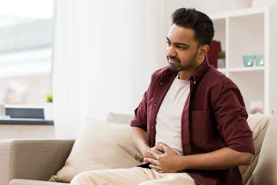A young man is seated on a couch. Both his hands are on his stomach and looks like he is nauseated and uncomfortable