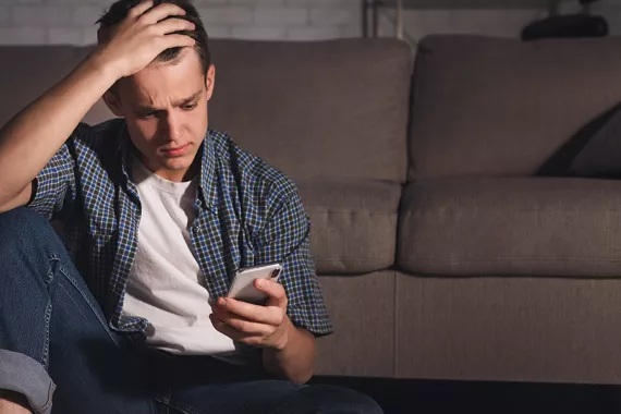 A teen boy sitting on the floor, leaning back against a couch. He looks worried as he gazes at his cellphone