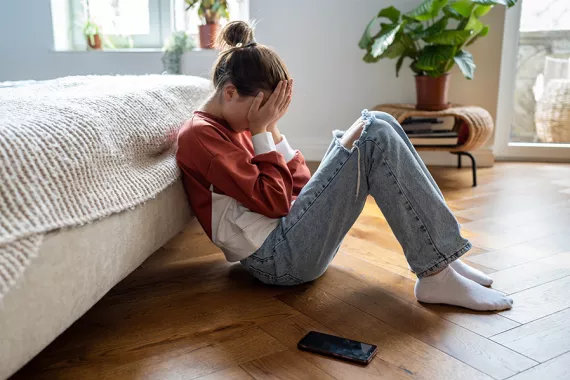 A young girl wearing jeans and a long-sleeve shirt sits on the floor holding her head in her hands. Her phone is on the floor near her.