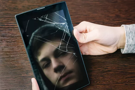 Two hands are holding a cellphone with cracked glass. The reflection in the glass shows the face a young teen boy wearing a hoodie