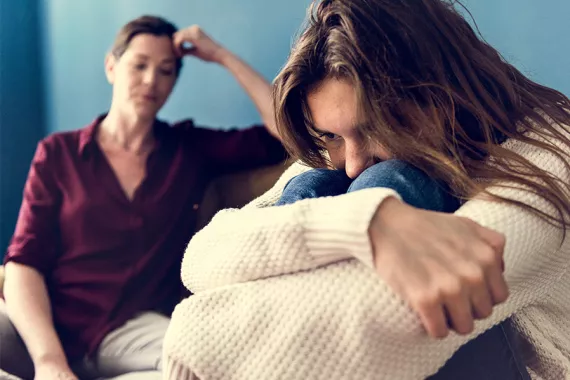 An anxious young woman sits in the fetal position while an older woman sitting in the background closes her eyes in frustration.