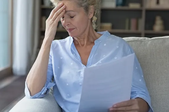 A woman is sitting on a couch and holding documents. Her eyes are closed and she is touching her forehead. She appears to be worried