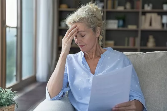 A woman is sitting on a couch and holding documents. Her eyes are closed and she is touching her forehead. She appears to be worried