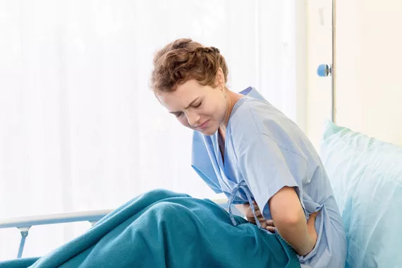 A young woman sitting in a hospital bed. She is leaning forward with her hands over her stomach. She is distressed and in pain