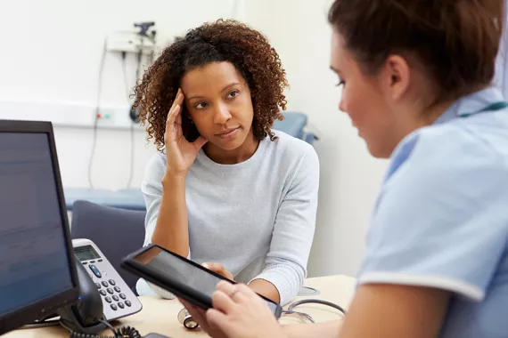 A young Black woman talks with a nurse in a clinical office setting. They are discussing information on an iPad and a desktop computer