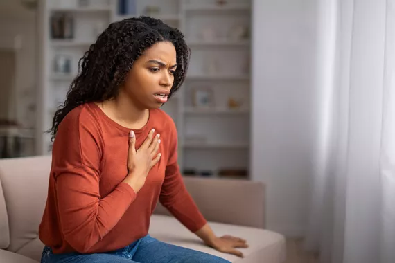 A young Black woman sits on a couch in a living room. She has her hand on her chest and appears worried