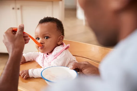 A man is feeding an infant some baby food with a spoon. They are sitting at a kitchen table and appear content and happy