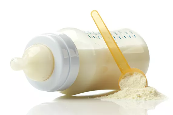 A baby bottle filled with formula lying on its side on a table. A small pile of loose formula with a measuring spoon is beside the bottle