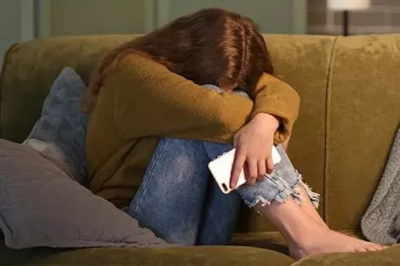 A teen girl curled up on a couch with her arms around her knees and her forehead on her knees. She holds a cellphone and looks distressed