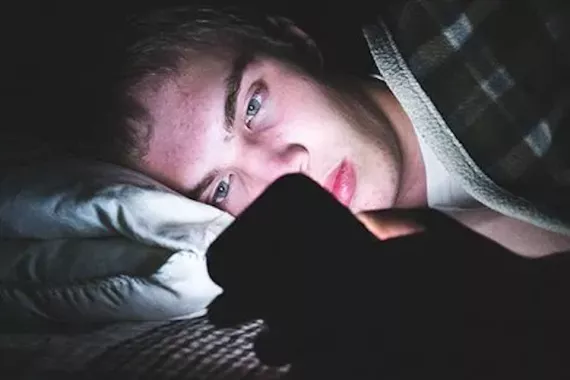 A young man lies on a bed and looks at his phone in the dark.